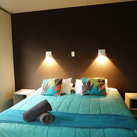 Taupo Debretts Spa Resort Екстер'єр фото The image shows a bedroom with a modern and inviting layout. The focal point is a bed with a vibrant turquoise comforter, decorated with two colorful pillows featuring floral patterns. On either side of the bed, there are small nightstands with wall-
