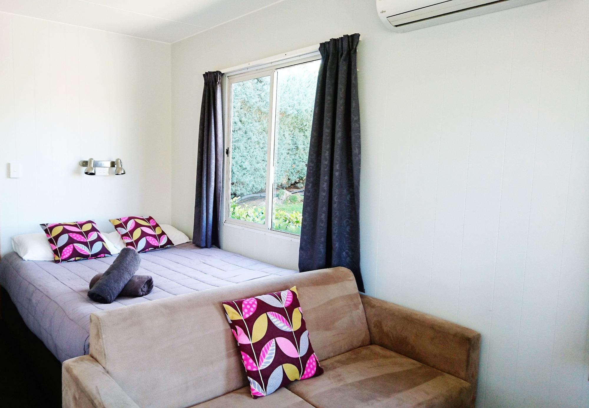 Taupo Debretts Spa Resort Екстер'єр фото The photo shows a cozy room with a bed and a small sofa. The bed features two decorative pillows with colorful patterns, and there is a rolled-up towel placed on the bed. Large windows are covered by dark curtains, allowing some natural light to ente