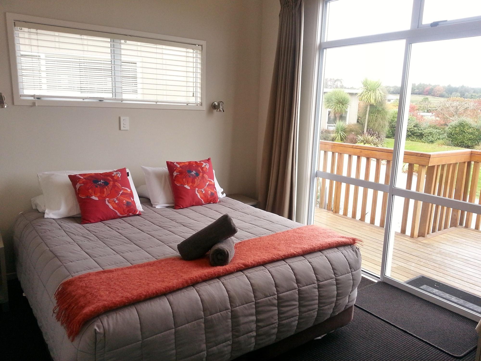 Taupo Debretts Spa Resort Екстер'єр фото The photo shows a cozy bedroom with a large bed centered in the room. The bed is covered with a light gray quilt and has decorative pillows in shades of red and orange. There is a bright orange throw blanket at the foot of the bed. A rolled towel is 
