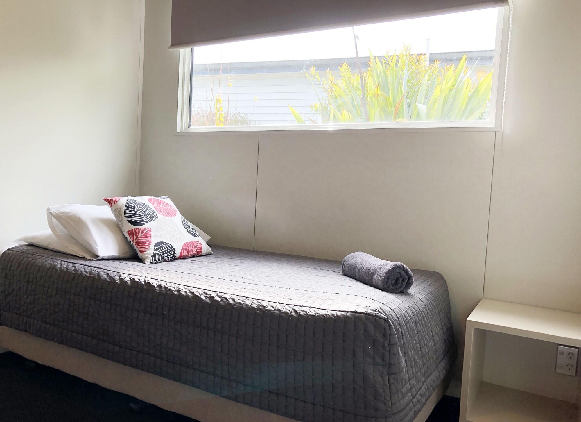 Taupo Debretts Spa Resort Екстер'єр фото The photo shows a simple bedroom. It features a single bed with a gray blanket, a couple of pillows (one patterned and one plain), and a rolled-up towel placed at the foot of the bed. Next to the bed is a small white nightstand. A large window lets i