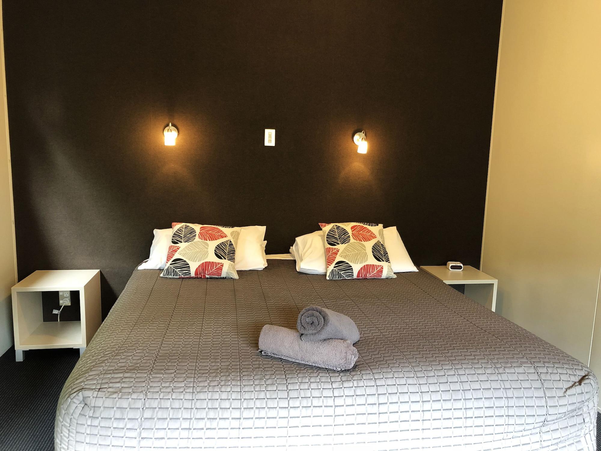 Taupo Debretts Spa Resort Екстер'єр фото The photo shows a bedroom with a large bed that has a gray quilted cover and decorative pillows. The walls behind the bed are painted a dark color, and there are two small wall-mounted lamps providing light. On either side of the bed, there are small