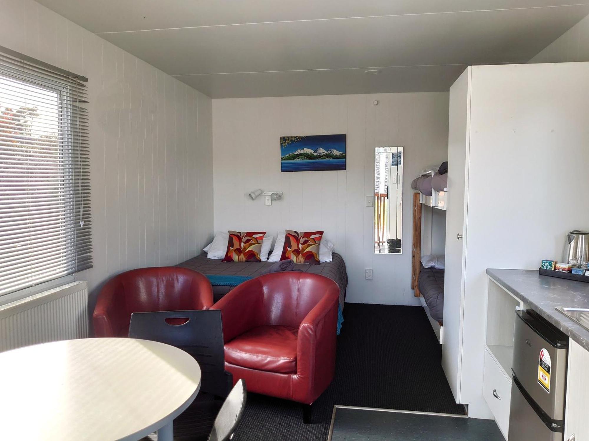 Taupo Debretts Spa Resort Екстер'єр фото The photo shows the interior of a small living space, possibly a cabin or studio apartment. There are two red armchairs and a round table in the foreground. The wall opposite has a bed with colorful pillows and a small nightstand. To the side, there 