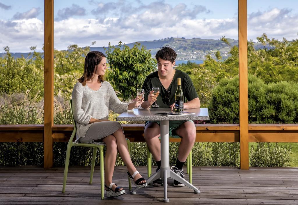Taupo Debretts Spa Resort Екстер'єр фото The photo shows a young couple sitting at a table outdoors. They are enjoying drinks, with the woman wearing a light-colored sweater and a skirt, while the man is dressed in a dark shirt and shorts. The table is set with two glasses and a bottle of w