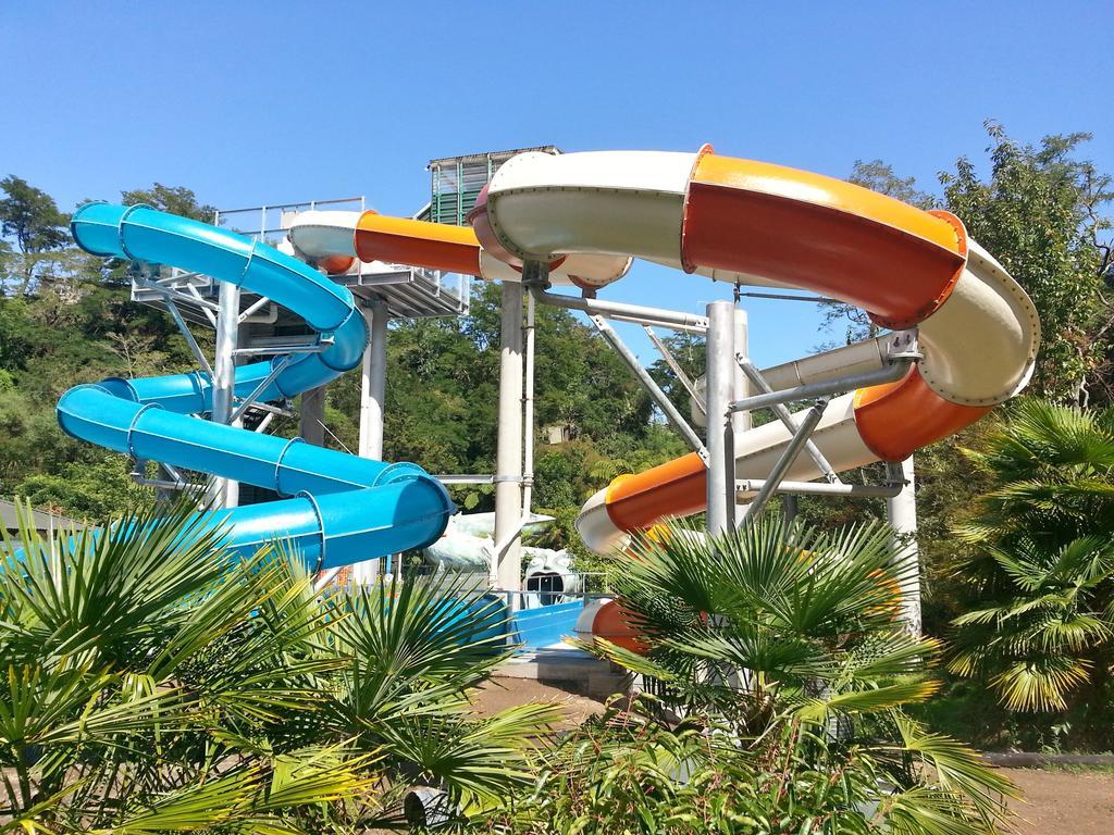 Taupo Debretts Spa Resort Екстер'єр фото The photo shows a water park featuring two large water slides. One slide is blue and curves gracefully, while the other is orange and white, twisting as it descends. They are supported by a metal structure and surrounded by lush greenery, including p