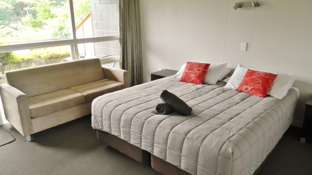 Taupo Debretts Spa Resort Екстер'єр фото The photo shows a cozy hotel or guest room. There is a large bed with gray bedding and two red decorative pillows. At the foot of the bed, there is a rolled-up towel. Next to the bed, there's a small bedside table and a light fixture on the wall. To 