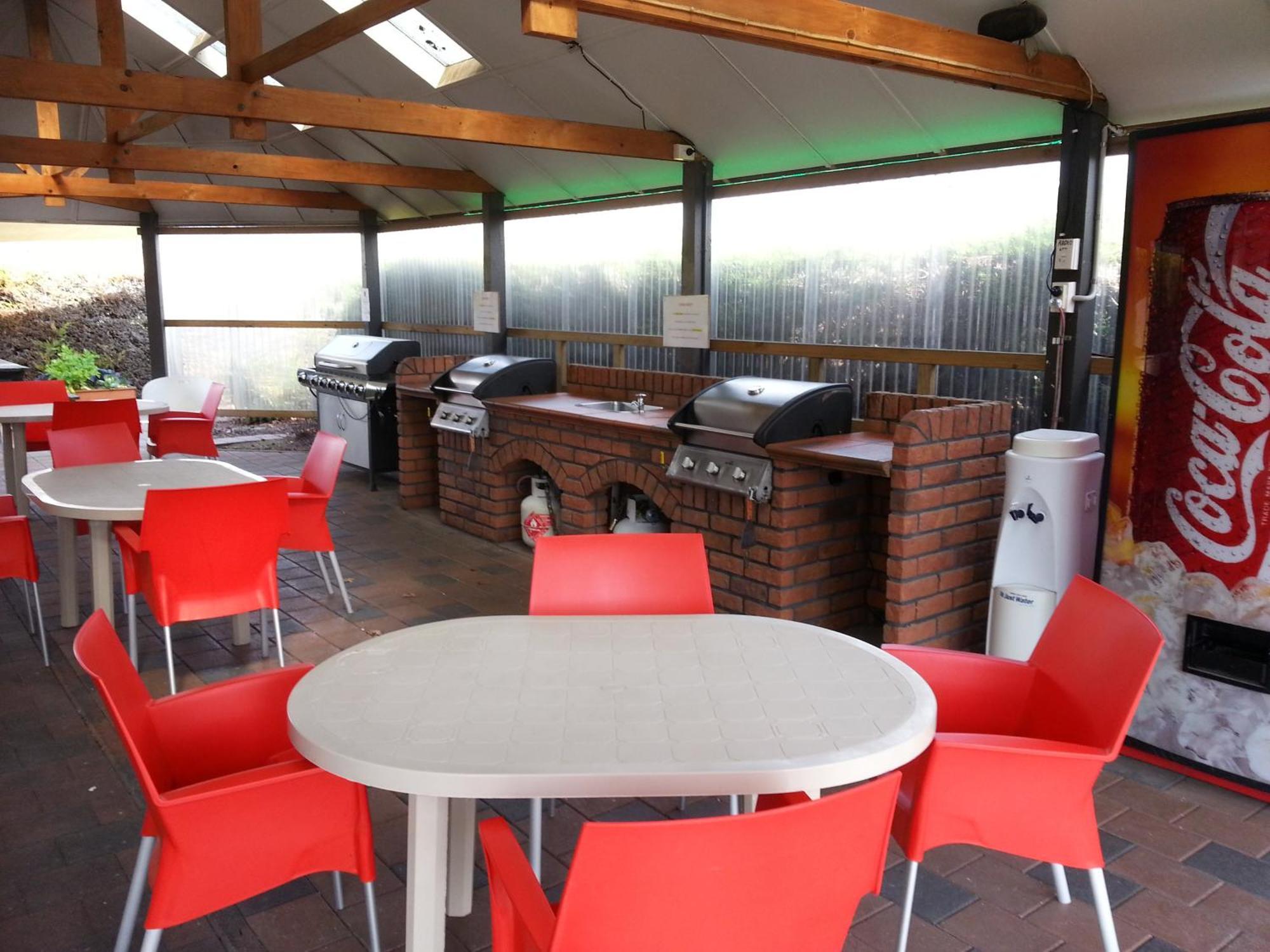 Taupo Debretts Spa Resort Екстер'єр фото The photo shows an outdoor kitchen area with a covered space. There are several barbecue grills set up on a brick counter. In the foreground, there are round tables with white tops and bright red chairs arranged around them. A Coca-Cola vending machi