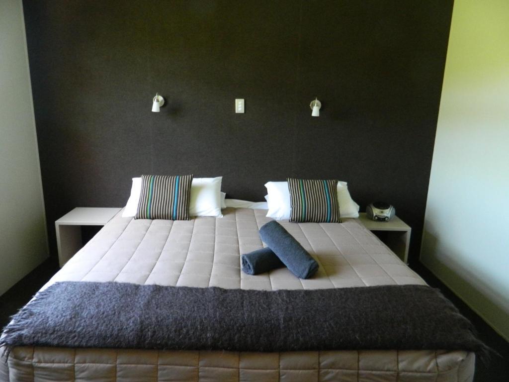 Taupo Debretts Spa Resort Екстер'єр фото The photo shows a modern bedroom featuring a large bed with a neutral-colored quilt and decorative pillows. There are two bedside tables on either side of the bed. The wall behind the bed is dark, creating a contrast with the lighter bedding. Additio