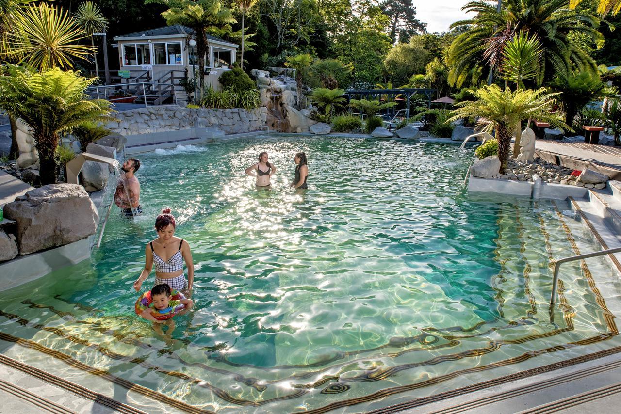 Taupo Debretts Spa Resort Екстер'єр фото The photo shows a scenic outdoor swimming pool surrounded by lush greenery, including palm trees. In the pool, several people are enjoying the water, with one woman holding a child who is in a flotation device. The atmosphere appears relaxed and invi