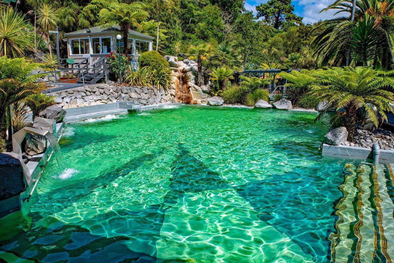 Taupo Debretts Spa Resort Екстер'єр фото The photo showcases a vibrant, natural-looking pool with crystal-clear, turquoise water. Surrounding the pool are lush green plants and palm trees, creating a tropical atmosphere. In the background, there is a white building, possibly a facility or a